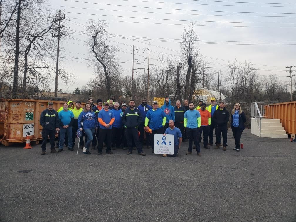 Volunteers in bright shirts pose outdoors near equipment; a “Colon Cancer Awareness” banner is in front.